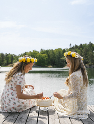 Två kvinnor med blomsterkransar sitter på en brygga och äter jordgubbstårta