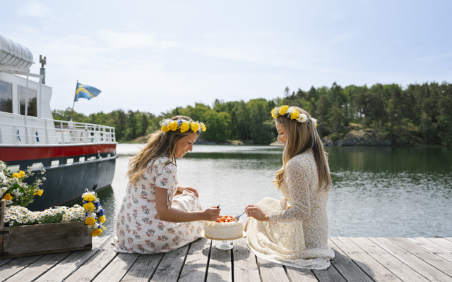 Två kvinnor med blomsterkransar sitter på en brygga och äter jordgubbstårta